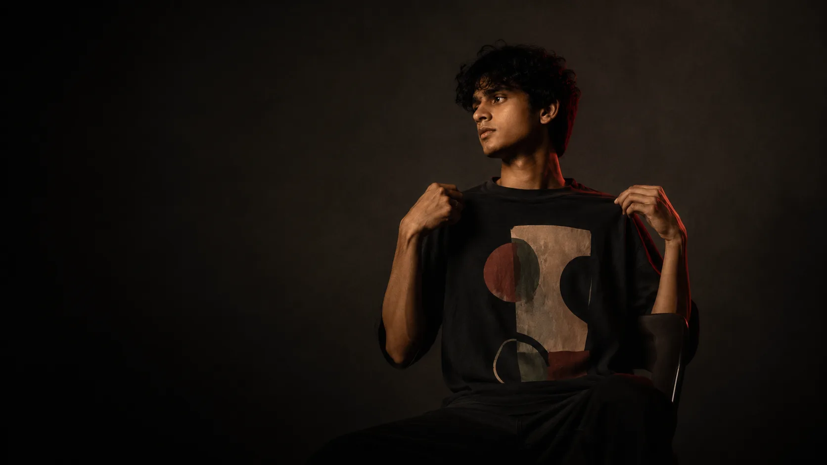 A young creator pulling on their custom-printed Shoonya t-shirt in a moody studio portrait